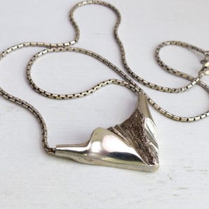 May include: A silver-toned necklace with a geometric pendant. The pendant has a textured, mountain-like design. The chain is a delicate, box-style link. The necklace is displayed on a white surface.