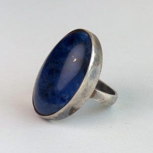 May include: A silver ring featuring a large, oval-shaped, deep blue cabochon stone. The metal band has a smooth, polished finish with slight signs of wear, and the stone displays a mottled texture with darker blue and black inclusions.