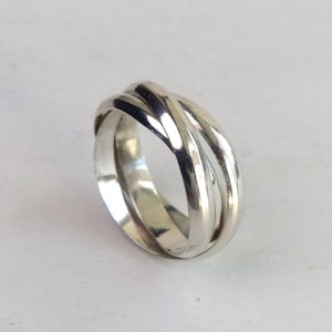 Può includere: Un anello in argento lucido composto da tre fasce intrecciate che si sovrappongono per creare una forma arrotondata e continua. La superficie metallica è liscia e riflettente, mostrando riflessi e ombre sugli anelli metallici curvi.