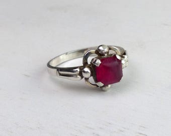 May include: A silver ring with a square, deep red gemstone. The ring has a decorative setting with small, round accents around the gemstone. The band is simple and polished, creating a classic and elegant look. Jewelry.