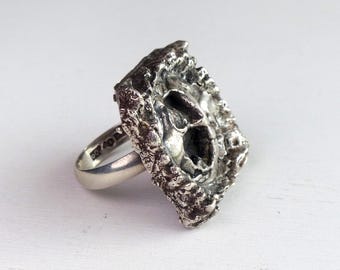 May include: A silver ring with a textured, rectangular face. The face has a rough, organic appearance with a central void. The ring band is smooth and polished. The ring is set against a white background.