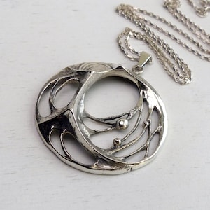 May include: A silver-toned pendant necklace with an abstract, openwork design. The pendant features a circular shape with intricate, flowing lines and small, spherical accents. The chain is also silver and lies next to the pendant on a white surface.