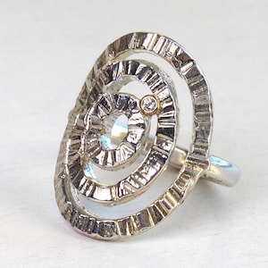 Op de afbeelding: Een zilverkleurige ring met een getextureerd, spiraalvormig ontwerp. De ring heeft een klein, rond, goudkleurig accent in het midden. De band is glad en gepolijst.