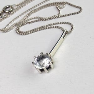 Könnte beinhalten: Eine silberfarbene Halskette mit einem dünnen, geraden Metallstab-Anhänger, der an einem runden, klaren, facettierten Stein in einer sechs-zackigen Fassung befestigt ist. Der Anhänger hängt an einer zarten, feingliedrigen silbernen Kette.