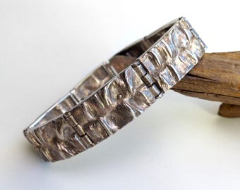 May include: A silver-toned bracelet with a textured, organic design. The bracelet is made of linked segments and has a weathered appearance. The bracelet is resting on a light background with a piece of wood.