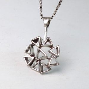 May include: A silver pendant necklace featuring a geometric design. The pendant is composed of multiple triangular shapes arranged in a circular pattern. The chain is a fine, silver box chain. The background is a light, neutral color.