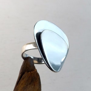 Könnte beinhalten: Ein silberner Ring mit einem großen, abstrakten, geschichteten Design. Der Ring hat eine polierte, reflektierende Oberfläche mit einer glatten, abgerundeten Form. Das Band ist schlicht und dünn, vor neutralem Hintergrund.
