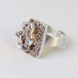 Könnte beinhalten: Ein silberner Ring mit einem strukturierten quadratischen Design. Der Ring hat eine strukturierte Oberfläche und ein leicht erhabenes Design.