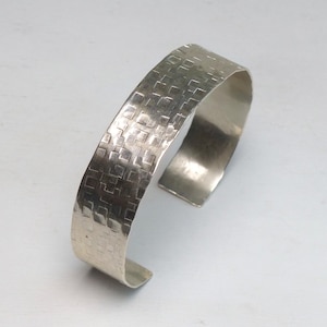 May include: A silver-toned cuff bracelet featuring a textured surface with a repeating pattern of small, recessed squares. The metal band has a polished finish and an open design, showing a smooth interior surface and slightly rounded edges.