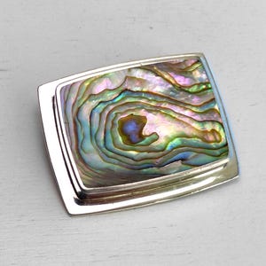 May include: A rectangular silver brooch with an iridescent abalone shell inlay. The shell displays swirling patterns of green, blue, and purple. The brooch has a polished silver frame and a secure pin backing. This jewelry piece is suitable for adding a touch of elegance.