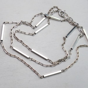 Può includere: Una collana color argento con un design a catena e accenti a barra rettangolare. La collana è disposta su una superficie di colore chiaro. La catena è composta da piccole maglie interconnesse.