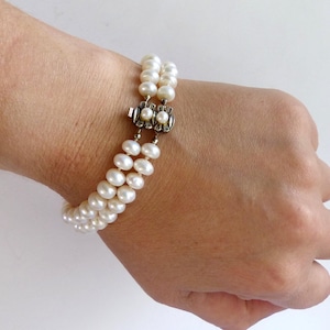 Könnte beinhalten: Ein doppelreihiges Perlenarmband mit einem silbernen Verschluss. Das Armband besteht aus zahlreichen cremeweißen Perlen, die zu zwei parallelen Reihen aufgereiht sind. Der Verschluss ist verziert und hat ein Blumenmuster. Das Armband wird am Handgelenk getragen.