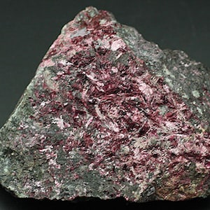 May include: A rough-hewn rock sample with a mix of gray and deep red hues. The red appears in a crystalline, layered formation across the surface of the stone. The rock has an irregular shape and a textured appearance.