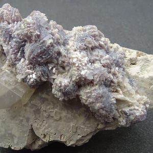 Puede incluir: Espécimen mineral natural con un grupo de cristales de color lavanda sobre una matriz gris claro. Una estructura cristalina clara es visible a la izquierda. La textura es áspera e irregular, destacando la belleza en bruto de la piedra.