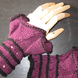 May include: A pair of knitted fingerless gloves in a deep purple hue, accented with black trim and stripes. The gloves feature a ruffled edge and are displayed on a wooden hand model, showcasing the intricate knit design.