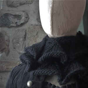 May include: A black knitted scarf with a textured, ruffled design. The scarf is draped around a mannequin's neck.