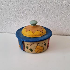 Op de afbeelding: Een kleine, ronde, keramische container met een deksel. De container is beschilderd met een kleurrijk, grillig ontwerp met geel, oranje, blauw, groen en rood. Het deksel heeft een lichtblauwe knop.