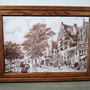 May include: A framed tile art piece depicting a sepia-toned European street scene. The artwork features a canal, buildings, trees, and people. The frame is made of dark brown wood.