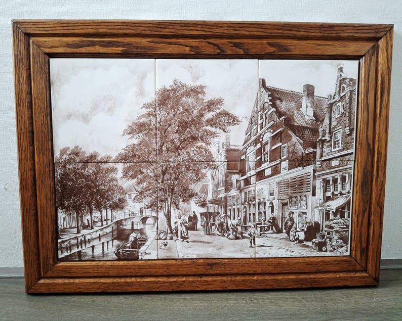 May include: A framed tile art piece depicting a sepia-toned European street scene. The artwork features a canal, buildings, trees, and people. The frame is made of dark brown wood.