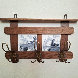 May include: Wooden wall shelf with two blue and white ceramic tiles depicting Dutch landscapes. The shelf has three brass hooks with decorative lion faces. The shelf is 30 cm wide.