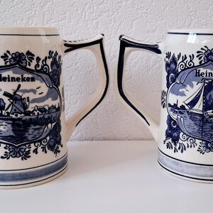 May include: Two blue and white ceramic beer steins with a Dutch theme. Each stein features a windmill and the word "Heineken" in a decorative banner.