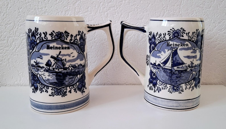 May include: Two blue and white ceramic beer steins with a Dutch theme. Each stein features a windmill and the word "Heineken" in a decorative banner.