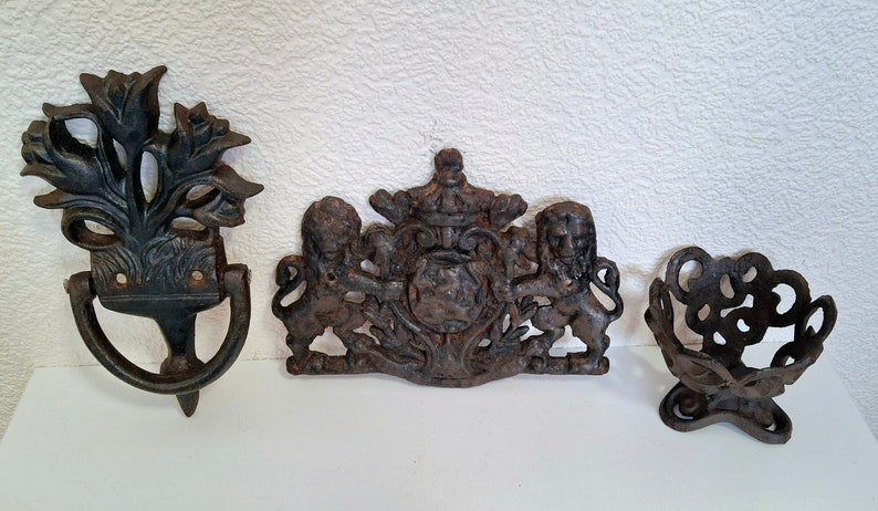 May include: Three antique cast iron door hardware pieces. A door knocker with a floral design, a decorative plaque with two lions and a crown, and a small decorative stand with a circular design.