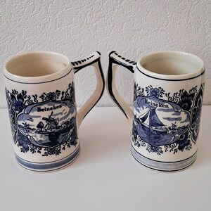 May include: Two blue and white ceramic beer steins with a Dutch design. The steins feature a windmill and a sailboat with the word "Heineken" on each.