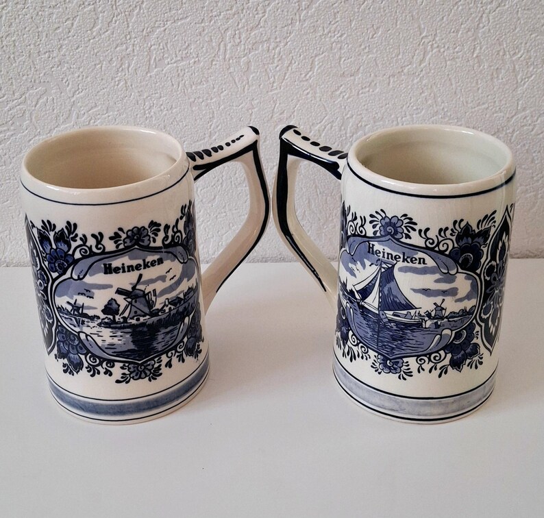 May include: Two blue and white ceramic beer steins with a Dutch design. The steins feature a windmill and a sailboat with the word "Heineken" on each.