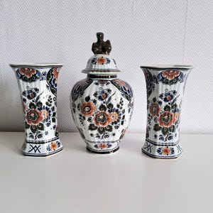 Może przedstawiać: Trzy ceramiczne wazony z kwiatowymi wzorami. Zestaw zawiera dwa pasujące wazony i słoik z pokrywką. Wazony i słoik mają białą podstawę z niebieskimi, pomarańczowymi i zielonymi kwiatowymi wzorami. Słoik ma gałkę w kształcie ptaka.