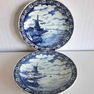 May include: Two blue and white decorative plates. Each plate features a windmill scene with a river and clouds. The plates have a scalloped edge and a detailed floral border. The plates are a classic design.