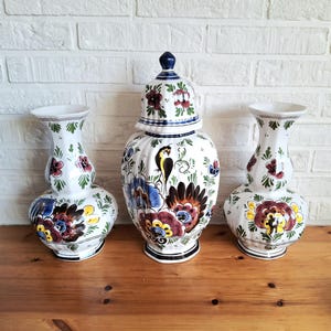 May include: Three white ceramic vases with floral and bird designs. The central vase has a lid. The vases feature a mix of red, blue, yellow, and green floral patterns. The vases are on a wooden surface.