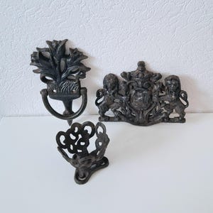 May include: Three black cast iron decorative wall hangings. One is a floral design with a ring, another is a crest with lions, and the third is a decorative basket.