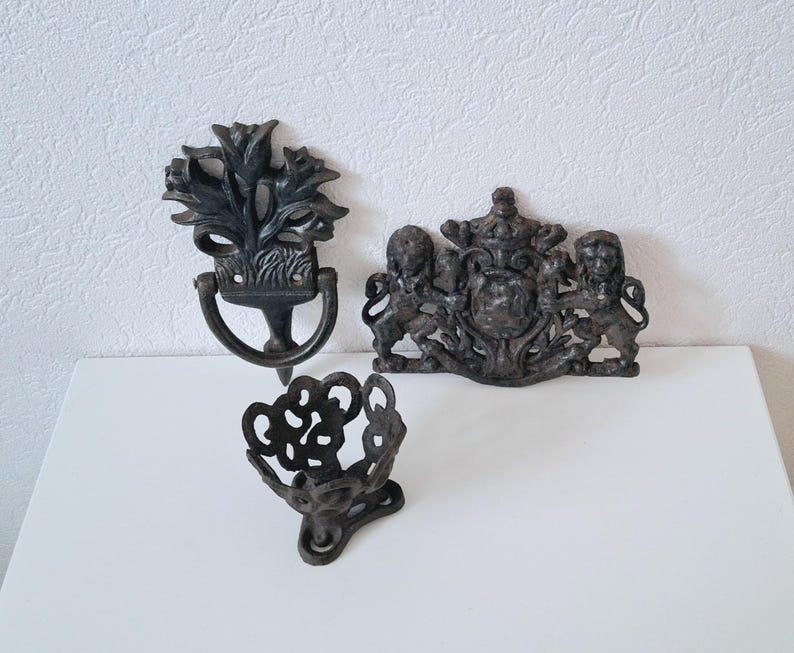 May include: Three black cast iron decorative wall hangings. One is a floral design with a ring, another is a crest with lions, and the third is a decorative basket.