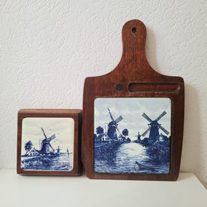 May include: Two wooden items with blue and white ceramic tiles featuring windmill scenes. One is a square coaster, the other a cutting board with a handle. The wood frames the tiles, creating a rustic, decorative look.