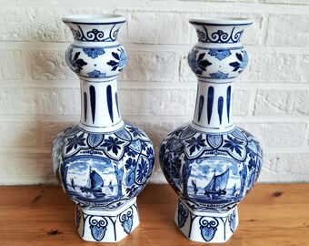 May include: Pair of blue and white ceramic vases. Each vase features a bulbous body with a detailed landscape scene, a slender neck, and a flared top. The design includes floral and geometric patterns, with a nautical theme.