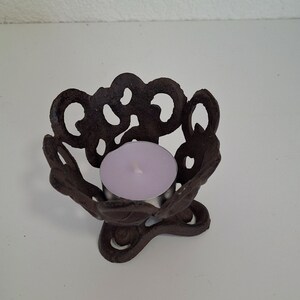 May include: A black metal candle holder with a decorative, intricate design. The holder has a round top with a small, round opening for a tea light candle. The holder has a three-legged base.