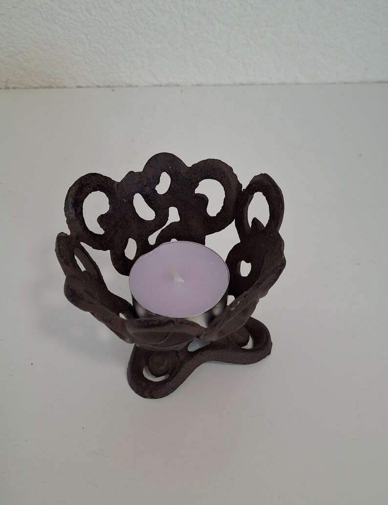 May include: A black metal candle holder with a decorative, intricate design. The holder has a round top with a small, round opening for a tea light candle. The holder has a three-legged base.