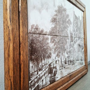 May include: A framed artwork featuring a sepia-toned depiction of a canal scene with trees, buildings, and a boat. The artwork is composed of multiple tiles and encased in a dark brown wooden frame.