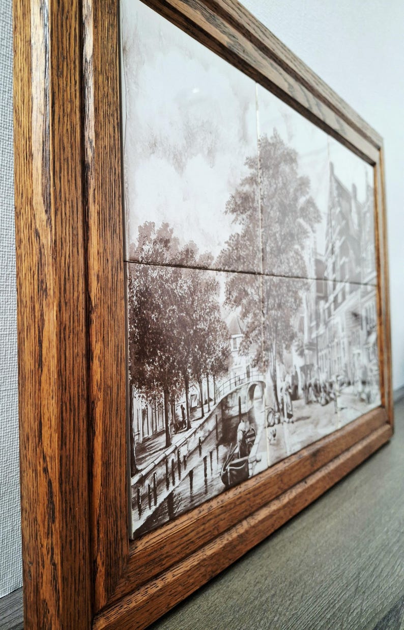 May include: A framed artwork featuring a sepia-toned depiction of a canal scene with trees, buildings, and a boat. The artwork is composed of multiple tiles and encased in a dark brown wooden frame.