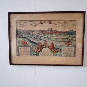 Peut inclure: Impression d'une carte ancienne encadrée, représentant une illustration détaillée et colorée d'une ville avec une rivière et un paysage. L'impression comprend du texte et des éléments décoratifs, dans un cadre en bois foncé. Une œuvre d'art historique.