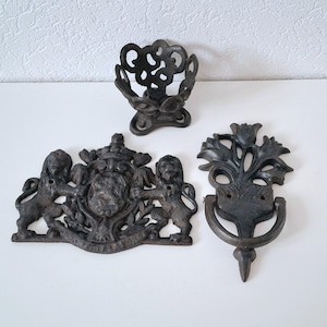 May include: Three black cast iron wall decor pieces. One is a decorative door knocker with a floral design, another is a coat hook with a lion design, and the last is a decorative ring.