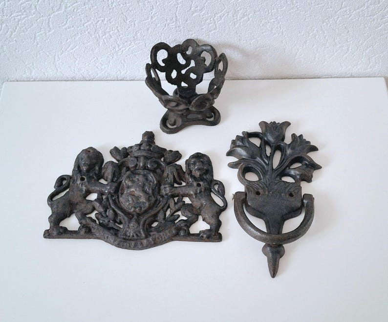 May include: Three black cast iron wall decor pieces. One is a decorative door knocker with a floral design, another is a coat hook with a lion design, and the last is a decorative ring.