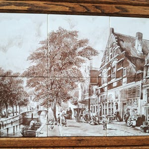 May include: A sepia-toned tile mural depicting a European town scene with a canal, bridge, and buildings. The artwork is framed in a dark wood. The scene includes people, trees, and boats, creating a vintage aesthetic.