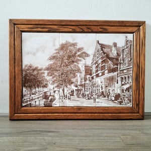 May include: A framed tile art piece depicting a sepia-toned European town scene. The artwork, composed of multiple tiles, showcases a canal, buildings, and people. The frame is made of dark wood.