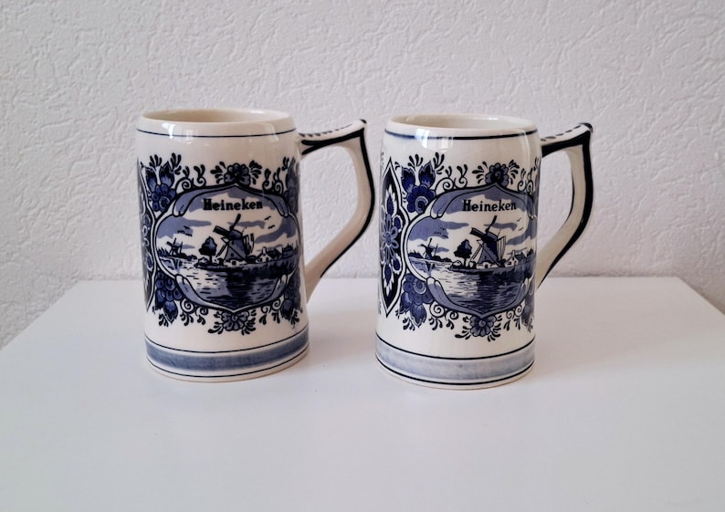 May include: Two white ceramic beer mugs with blue and white Delftware designs. The mugs feature a windmill and the word "Heineken".