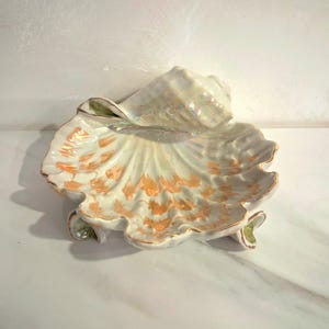 Puede incluir: Un plato de cerámica con forma de concha marina y acabado nacarado. El plato presenta un borde festoneado y está adornado con detalles en naranja y dorado. El plato está diseñado para parecerse a una concha.