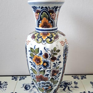 Może przedstawiać: Biały ceramiczny wazon z rozszerzaną górą i pękatym korpusem. Wazon zdobiony jest kwiatowym wzorem w odcieniach pomarańczu, błękitu i brązu, z zielonymi liśćmi. Wazon ma około 30 cm wysokości.