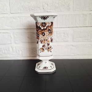 May include: A white ceramic vase with a floral design in shades of brown. The vase has a square top and base, with a cylindrical body. The floral pattern is detailed and covers the majority of the vase's body.