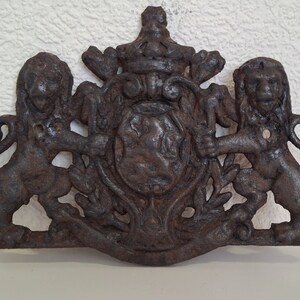 May include: A cast iron decorative plaque featuring two lions flanking a shield with a crown above. The plaque is in a dark brown color and has a distressed finish.